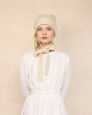 Woman wearing a white dress and beige bonnet with a bow against a beige background